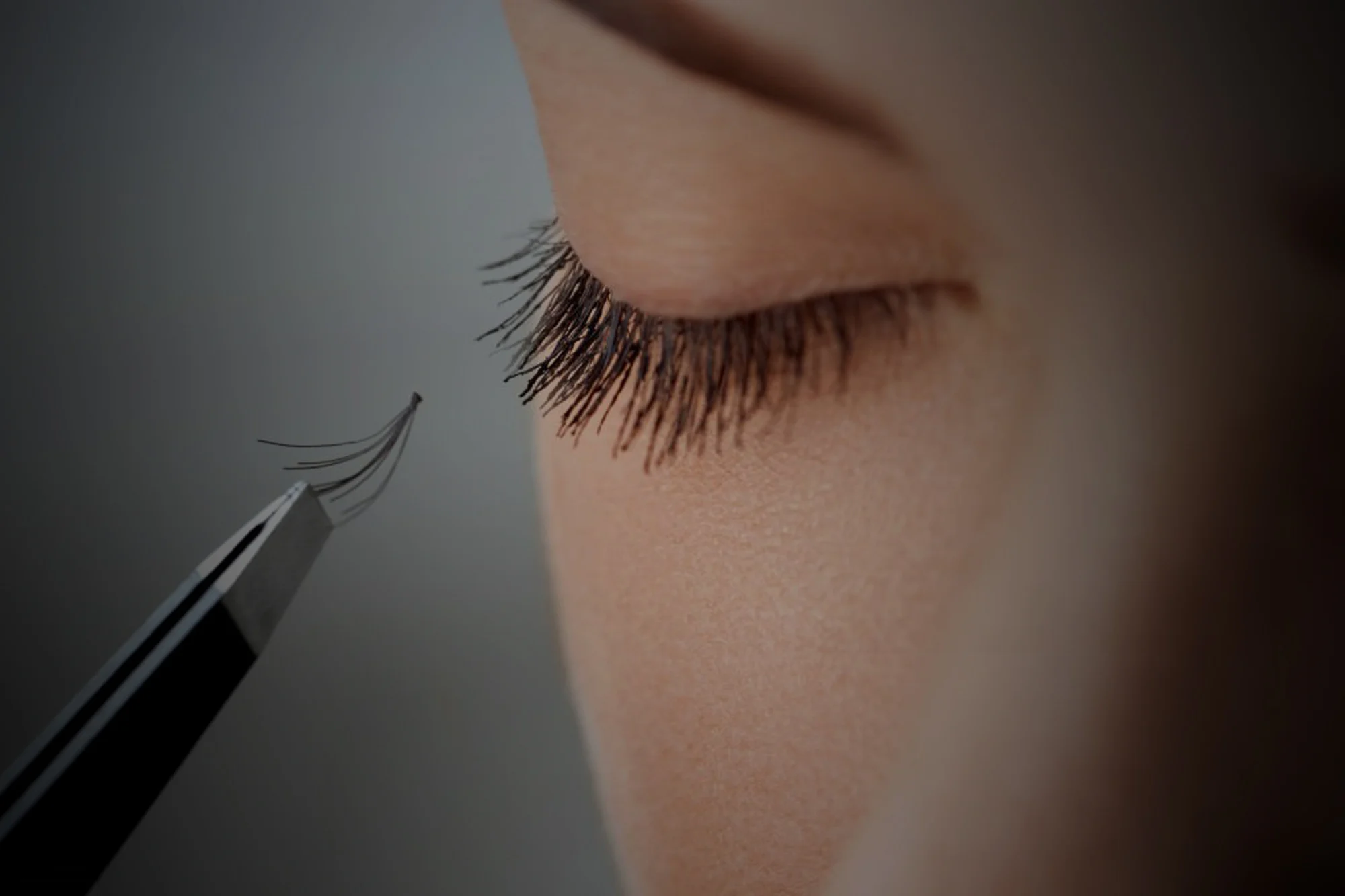 A woman with eyes closed and a tweezer with a fan of eyelashes being applied.